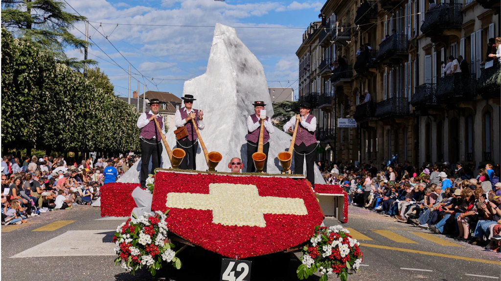 Flower-covered float with Swiss cross and alphorn players parading during the Neuchâtel Wine Festival, with mountain backdrop.