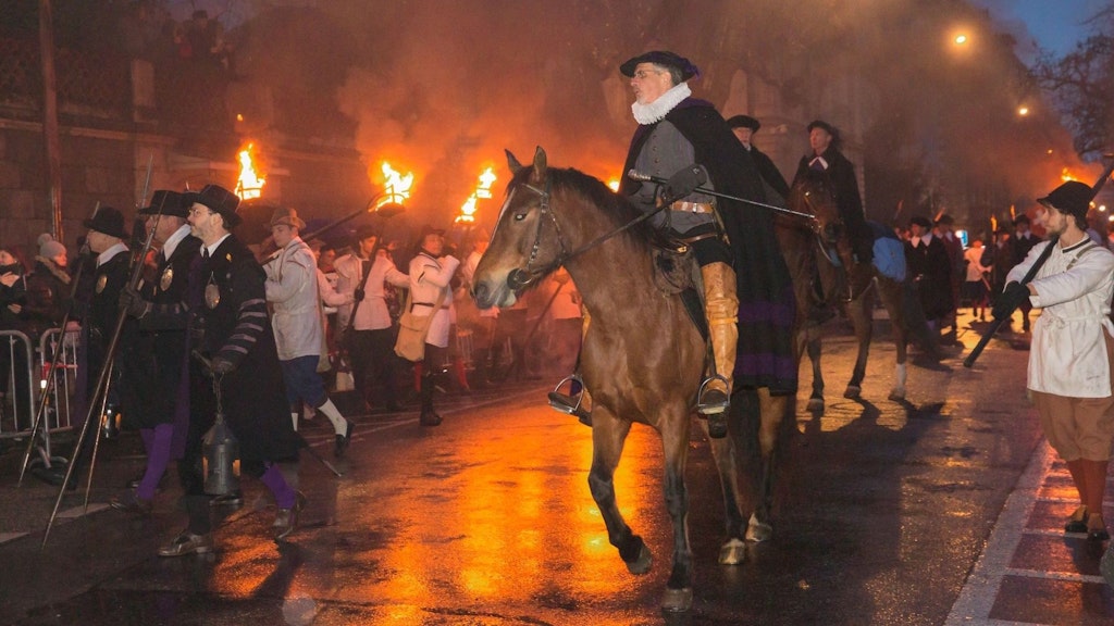 Nighttime parade with riders in historical costumes and torchbearers on a wet street.