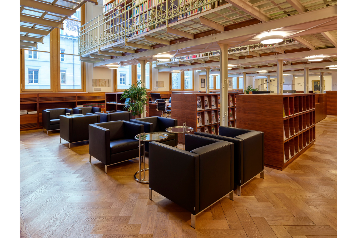 Modern reading area with black leather armchairs, round tables, magazine shelves, mezzanine library, parquet floor and large windows.