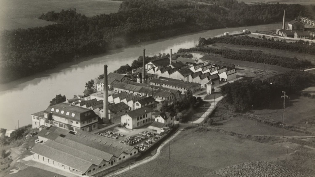 Black-and-white aerial view of a chemical plant by a river, showing multiple buildings, an access road and surrounding wooded areas. 