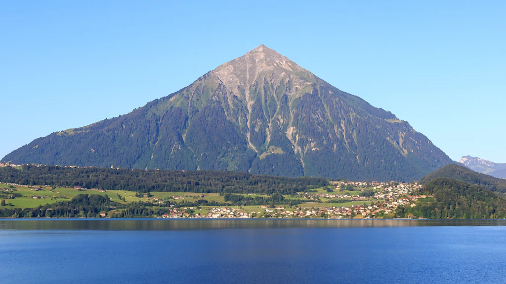 View of the pyramid-shaped Niesen with rocky summit, forested slopes and the lake in the foreground under a clear sky.