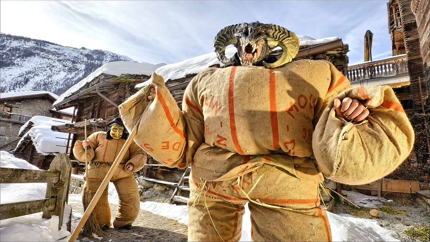 Dos personas con trajes rellenos de heno y máscaras, una de ellas con una cabeza de monstruo con cuernos, desfilan por una calle nevada de un pueblo alpino.
