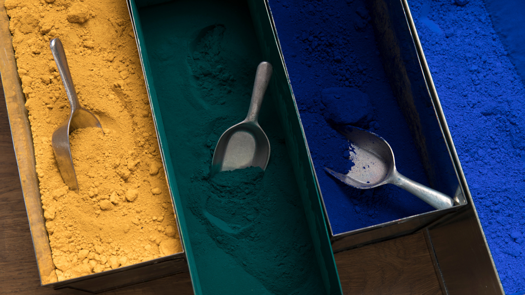 Tres bandejas metálicas en fila con polvo pigmentado (ocre amarillo, verde oscuro, azul intenso) y una pala de metal parcialmente enterrada.