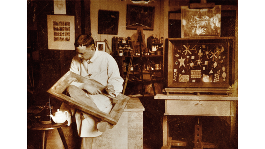 Fotografía histórica de un hombre sentado en un taller de arte con un marco en las manos, con pinturas, caballetes y lienzos al fondo.