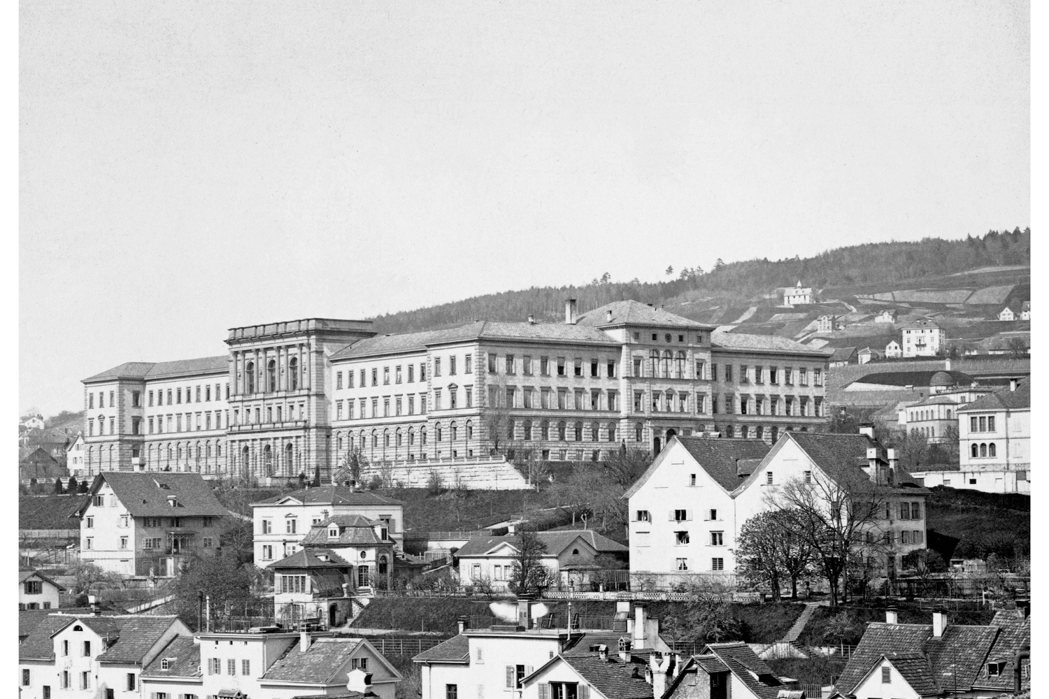 Vista histórica en blanco y negro del edificio principal original de la ETH Zúrich, diseñado por Gottfried Semper en 1880, encaramado en una colina ante casas.