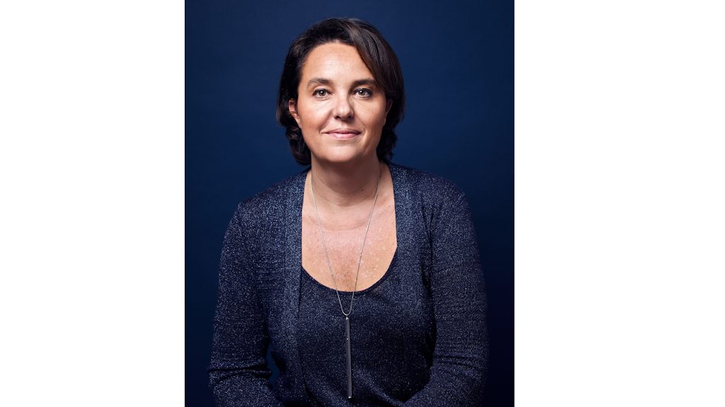 Retrato de una mujer de cabello castaño, con blusa azul brillante y un collar plateado largo, posando ante un fondo azul liso. 