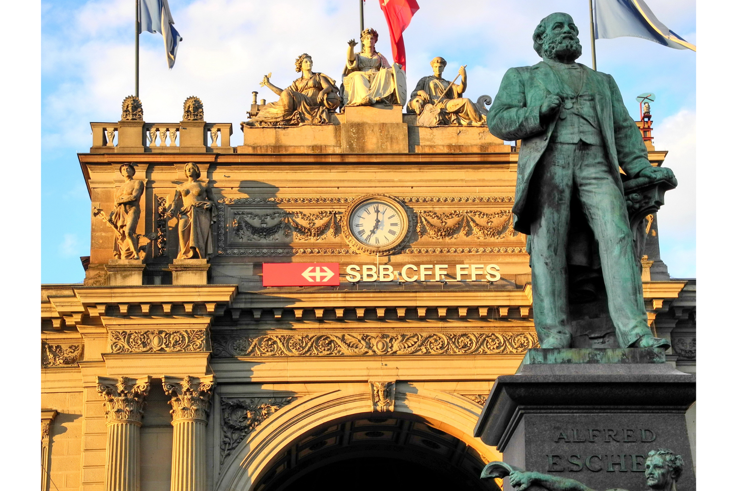 Estatua de bronce de Alfred Escher frente a la ornamentada fachada de la estación de Zúrich, con reloj, esculturas alegóricas y letrero «SBB CFF FFS» sobre el arco.