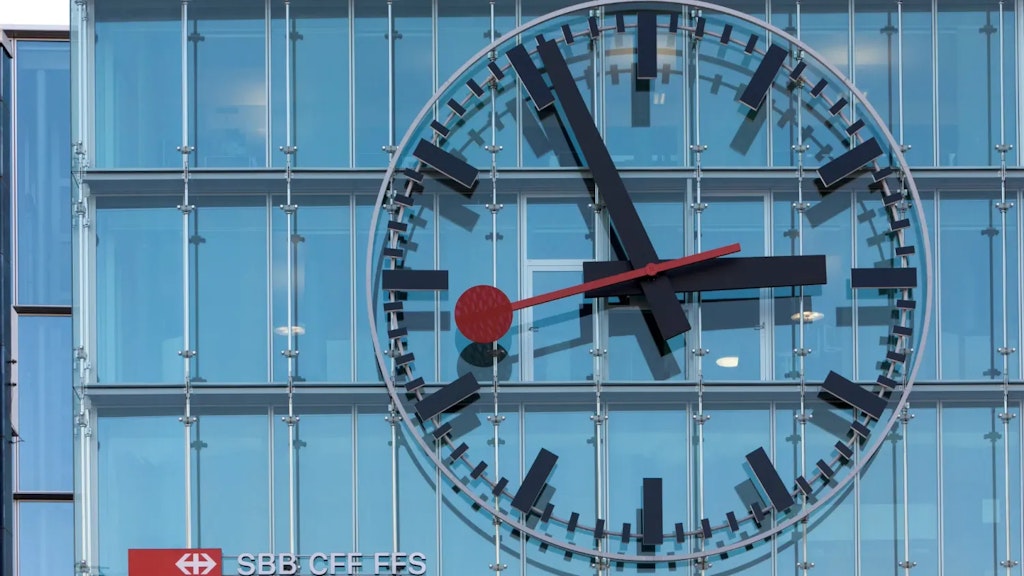 Gran reloj de estación suizo en una fachada de vidrio, con agujas negras y segundero rojo sobre el logotipo «SBB CFF FFS».