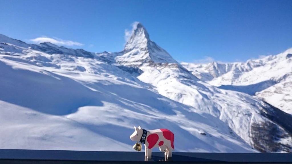 Una pequeña vaca de juguete está frente a un amplio paisaje montañoso cubierto de nieve con un pico destacado al fondo.