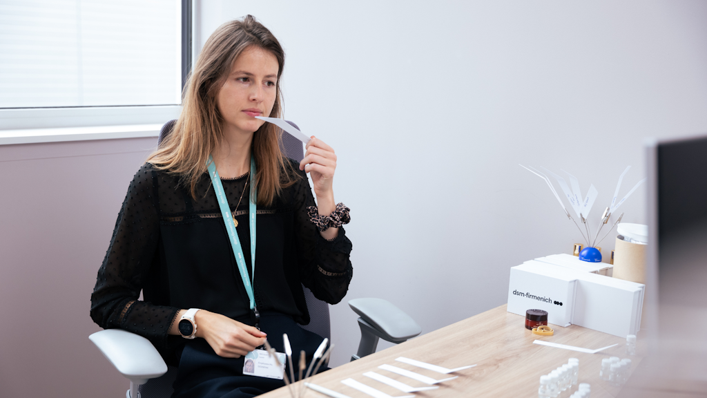 Una mujer sentada en un escritorio huele una tira de perfume, rodeada de tiras olfativas y muestras en un entorno de trabajo neutro.