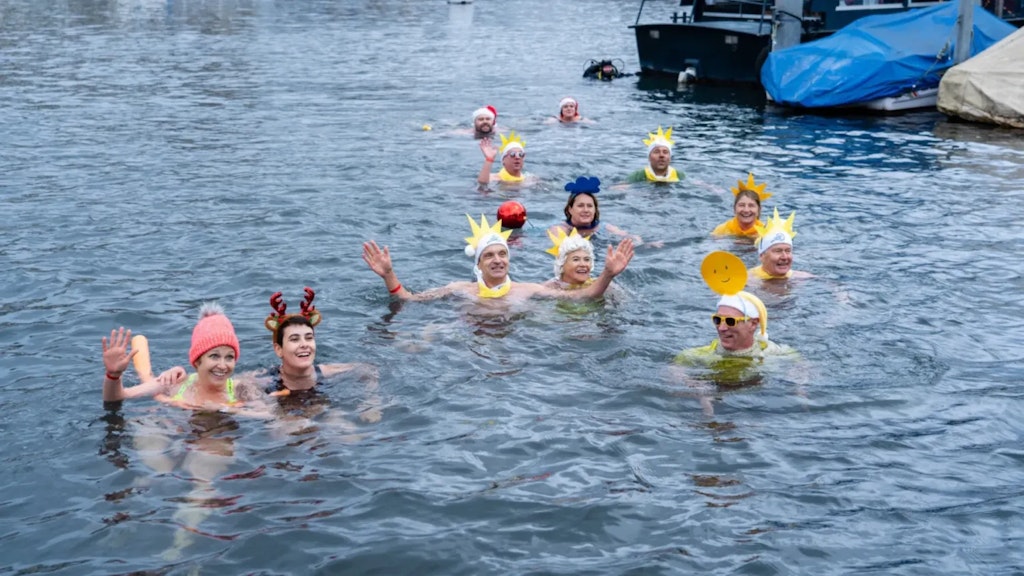 Grupo de personas nadando en agua fría con gorros coloridos y accesorios festivos, sonriendo y saludando.