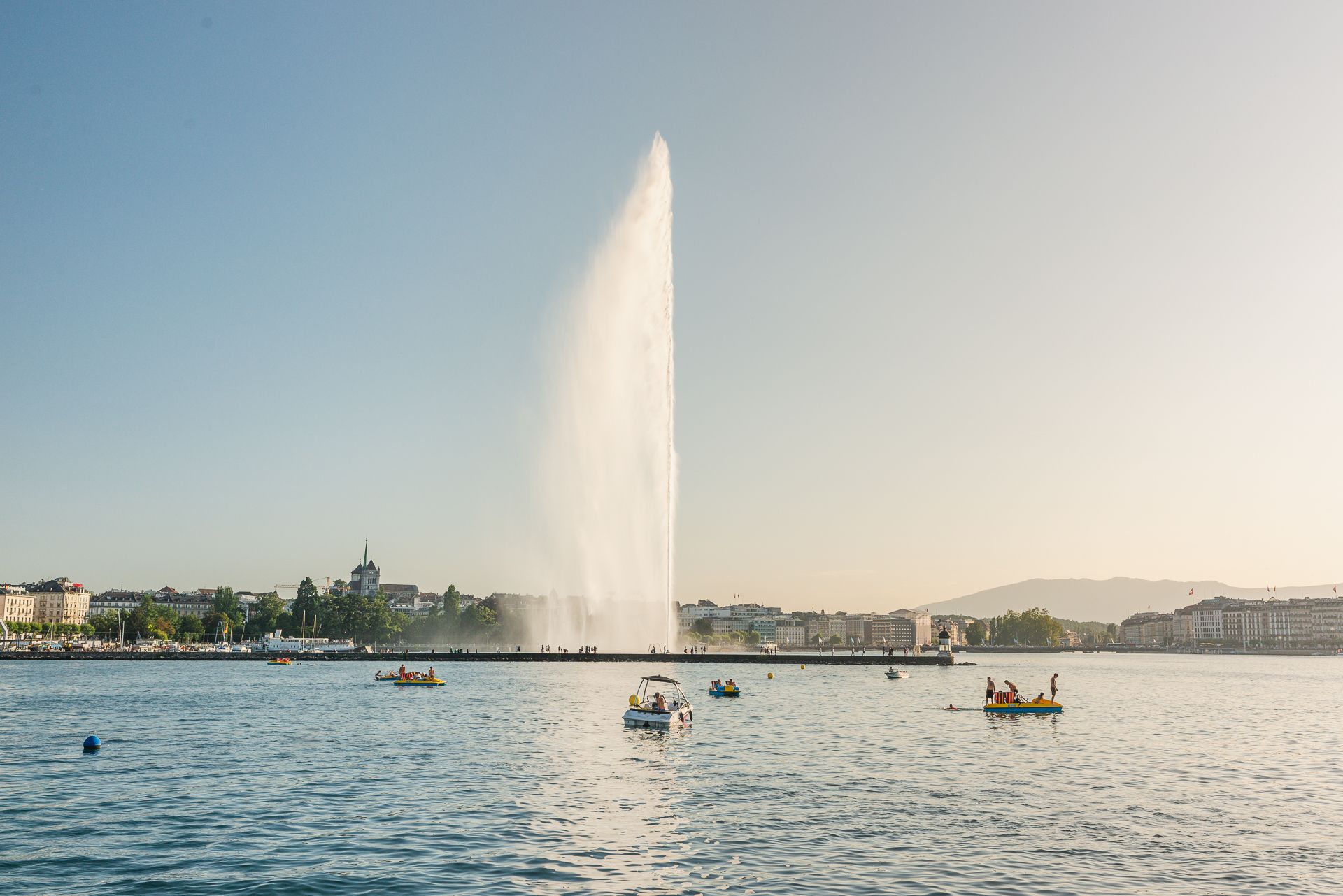 Vista de un lago con botes y el chorro del Jet d’Eau en el centro, edificios en la orilla y cielo despejado.