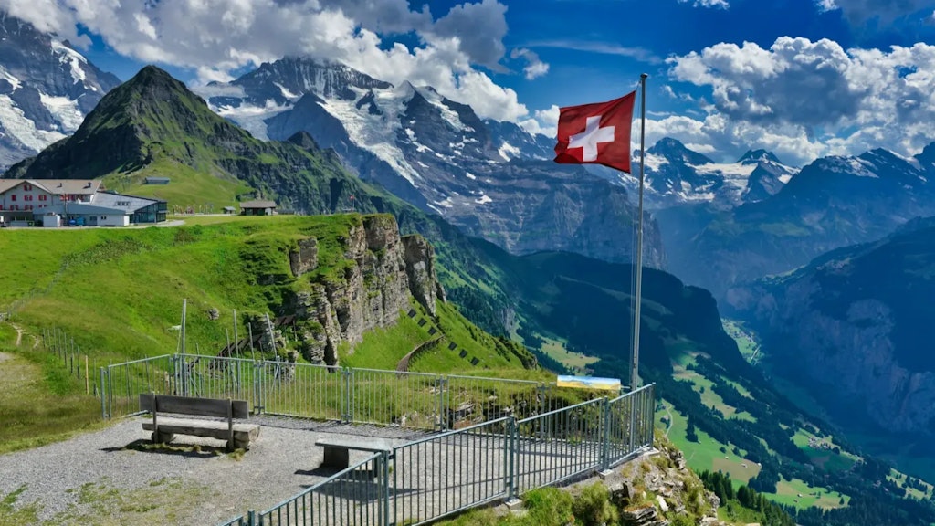 Bandera suiza roja con cruz blanca ondeando en un mirador con montañas nevadas y valles verdes bajo un cielo azul.