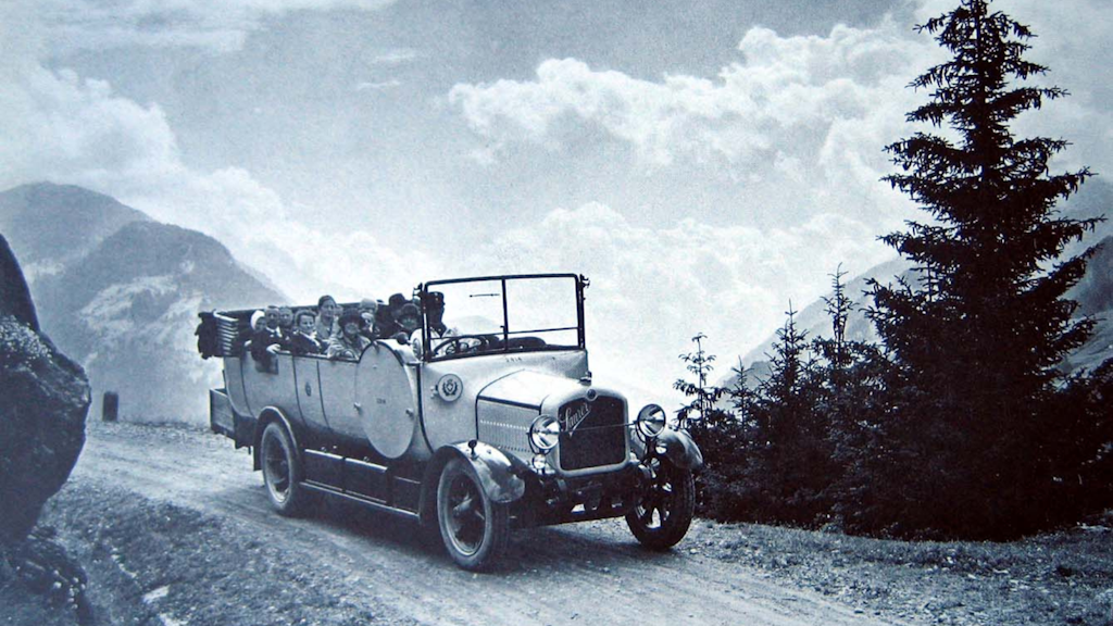 Autobús postal Saurer AD descubierto con pasajeros en una carretera de montaña en el puerto de San Gotardo, rodeado de árboles y picos rocosos bajo cielo nublado.