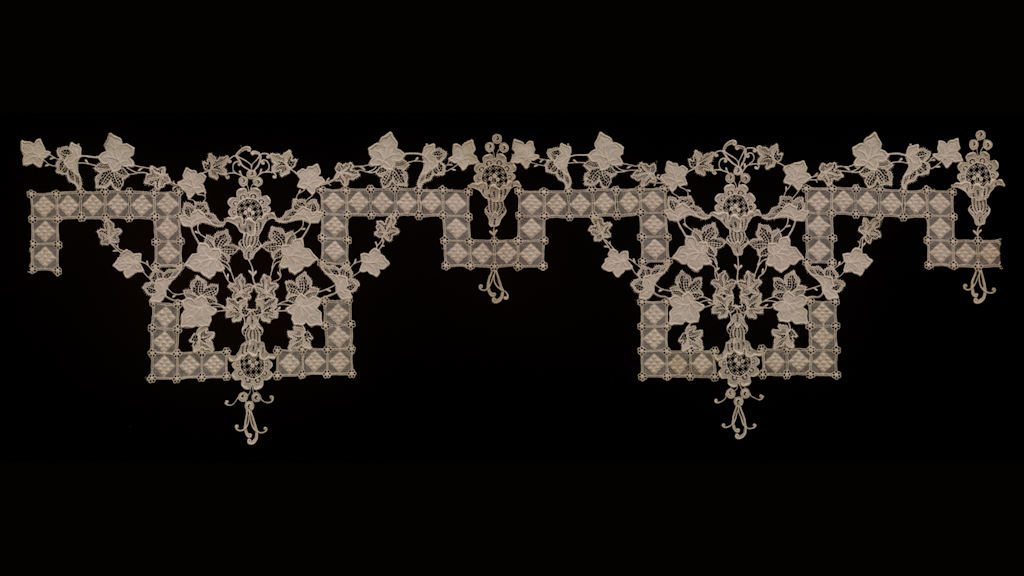 Muestra de encaje o bordado blanco con motivos florales y geométricos dispuestos simétricamente sobre fondo negro.