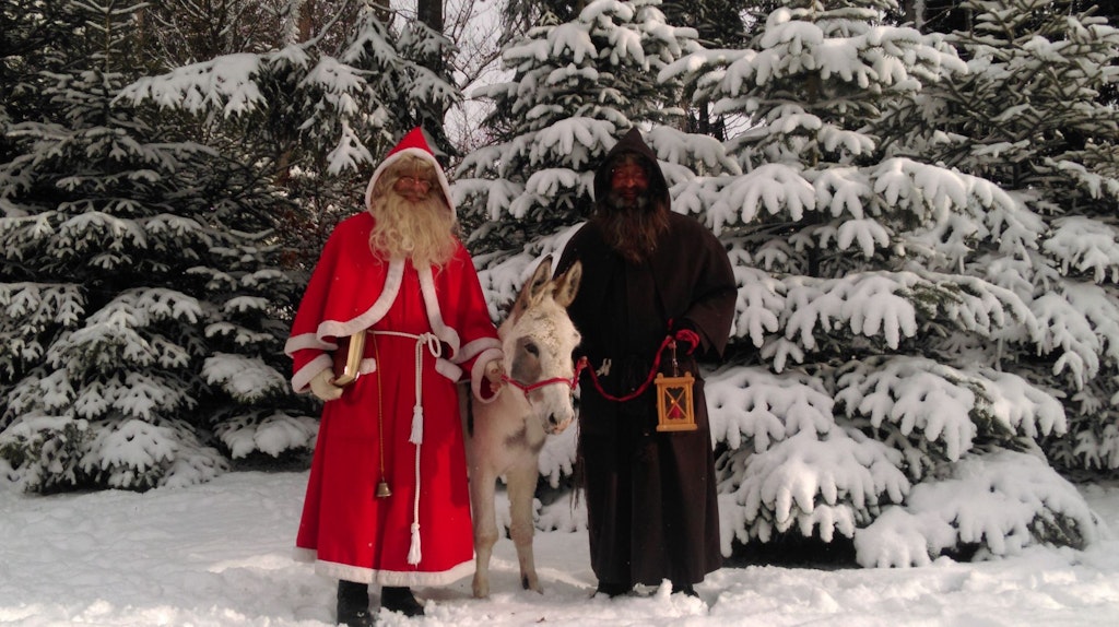 En un bosque nevado, dos hombres con largas barbas están de pie, uno lleva un abrigo rojo, el otro una túnica marrón, y entre ellos hay un burro gris.