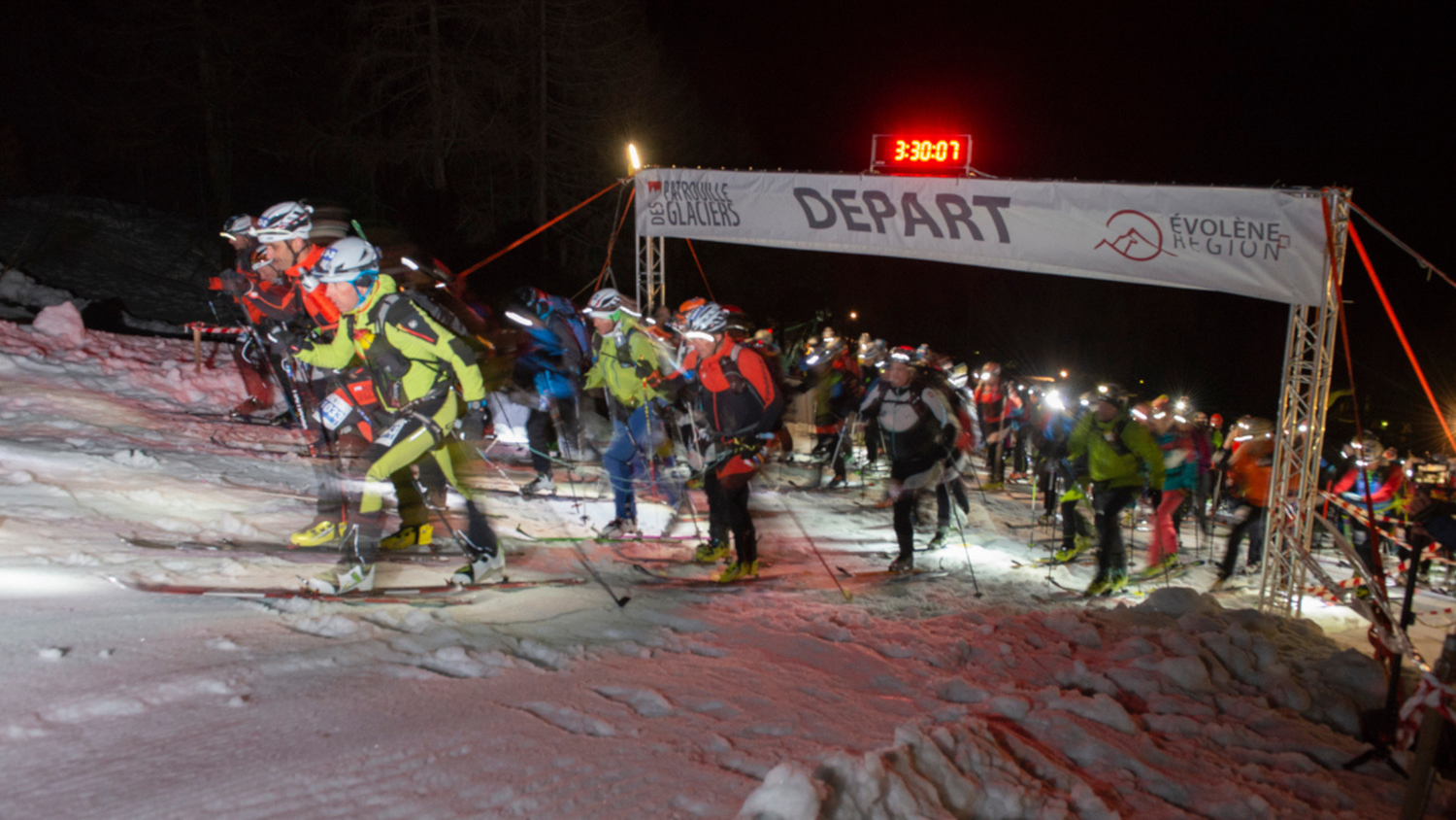 Salida nocturna de la carrera A1 desde Arolla a las 3h30, los esquiadores con linternas frontales comienzan bajo un arco «Départ».