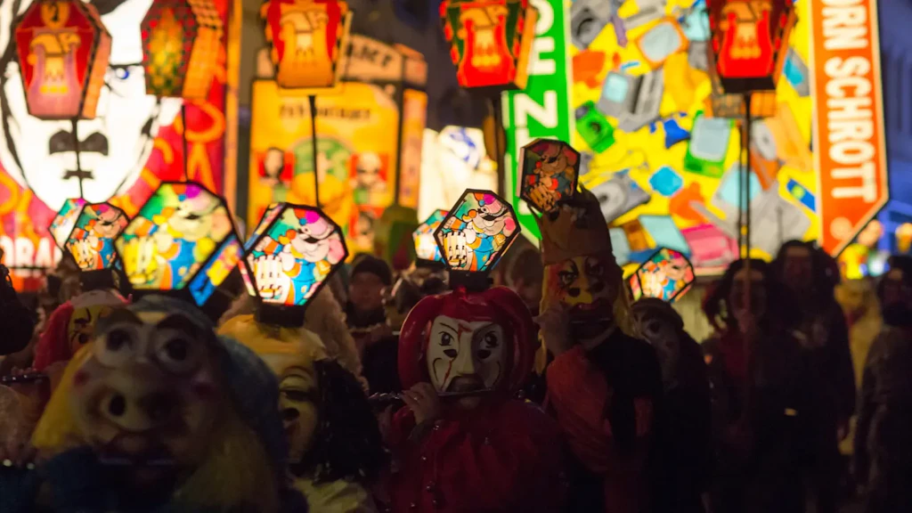 Desfile nocturno con personas enmascaradas portando linternas iluminadas, frente a paneles coloridos y una multitud en una calle urbana.