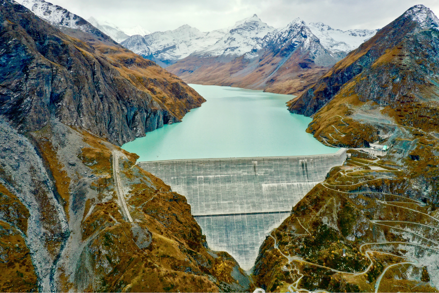 Vista aérea de un gran embalse de hormigón que retiene un lago turquesa entre montañas escarpadas parcialmente nevadas.