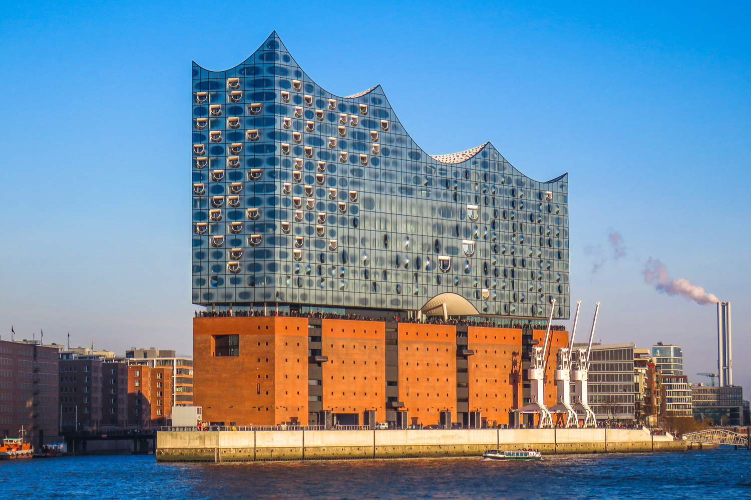 Vista exterior de la Elbphilharmonie en Hamburgo, edificio moderno de cristal sobre antiguo almacén de ladrillo, junto al río Elba en un día soleado.