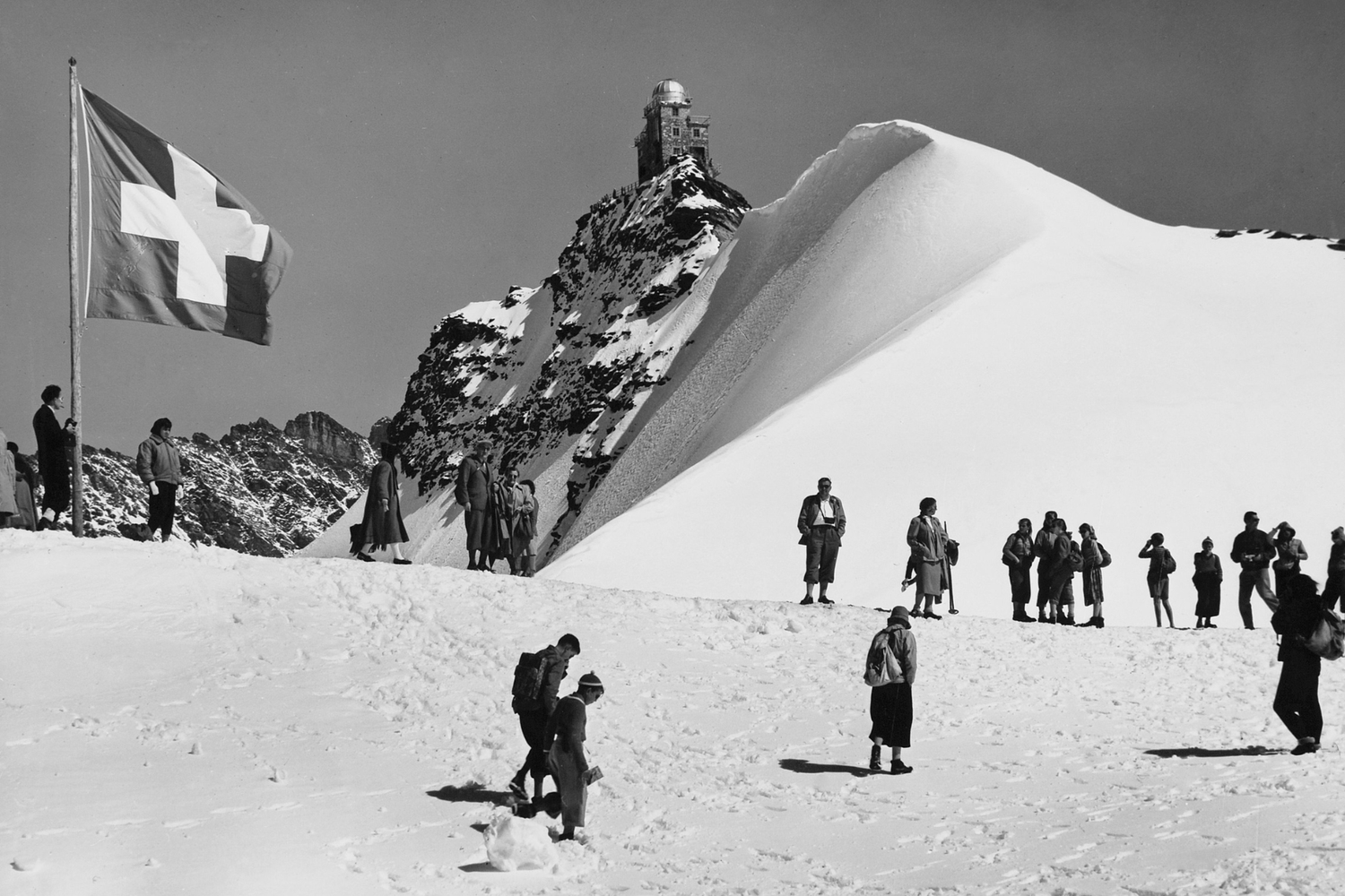 Personas caminando sobre la nieve cerca de una bandera suiza, con un observatorio en la cima de una montaña nevada al fondo.