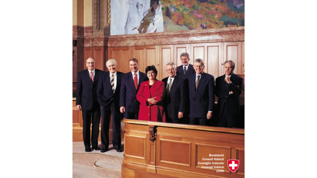 Foto oficial de 1999 del Consejo Federal suizo. Sus miembros y el Canciller posan de pie en una sala con revestimiento de madera.