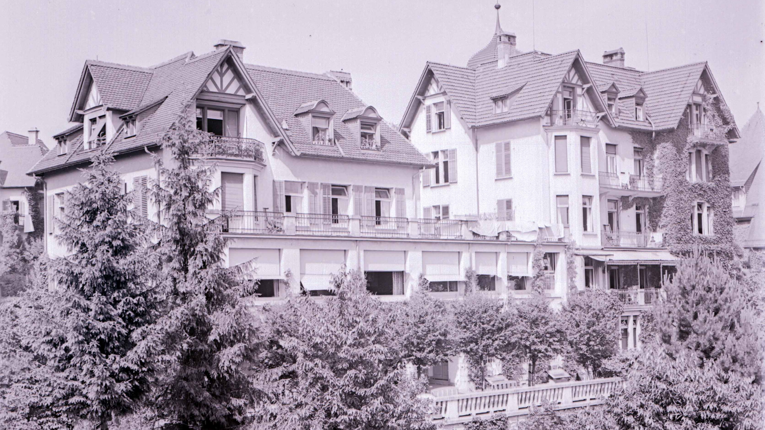 Fotografía en blanco y negro de un gran edificio de varias plantas con tejados inclinados, balcones y vegetación circundante, probablemente tomada en la década de 1920.