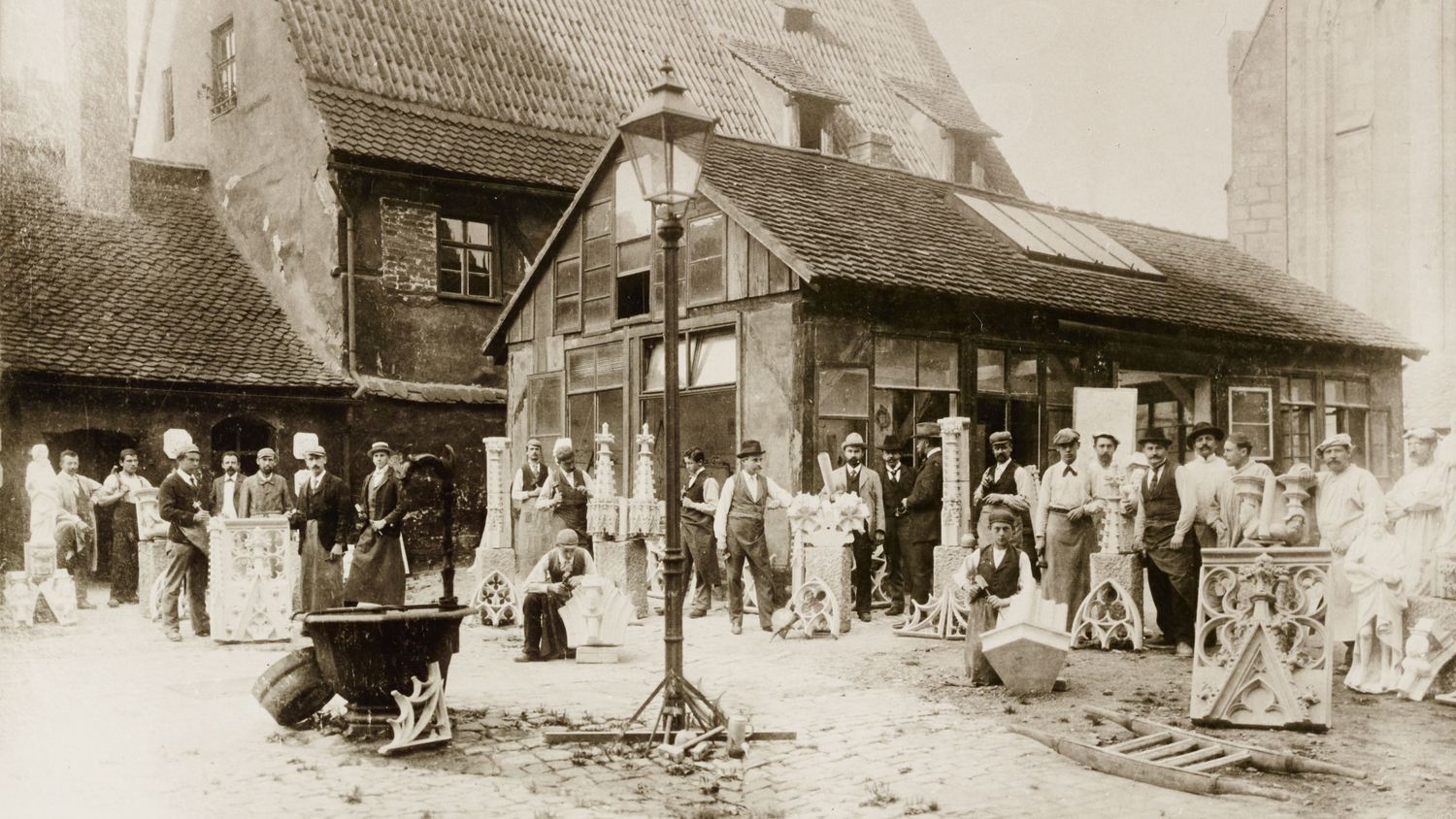 Fotografía histórica de artesanos de cantería trabajando en un patio pavimentado cerca de la iglesia de San Lorenzo en Núremberg, rodeados de elementos arquitectónicos esculpidos.