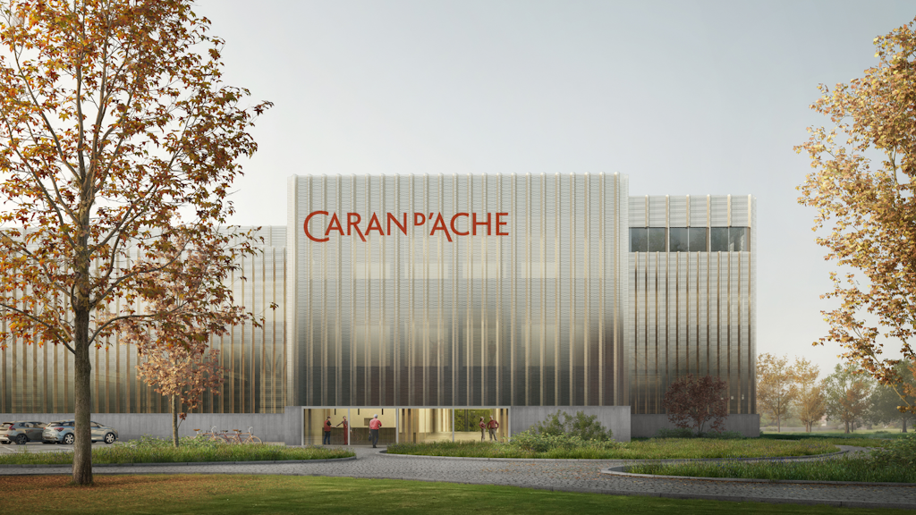 Fachada de un edificio moderno con logo Caran d’Ache, revestimiento metálico translúcido, entrada acristalada, árboles en primer plano y cielo claro. 