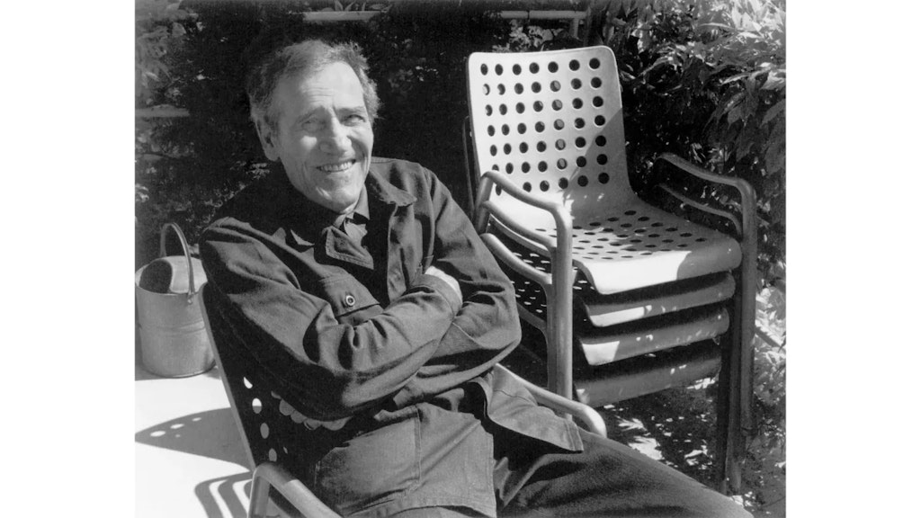 Hombre sentado al aire libre en una silla con los brazos cruzados; a su lado hay una pila de sillas metálicas perforadas, rodeadas de vegetación.