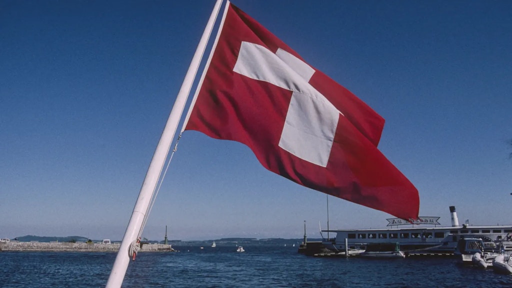 Bandera suiza roja con cruz blanca ondeando en un mástil sobre una masa de agua, con barcos e instalaciones portuarias al fondo.