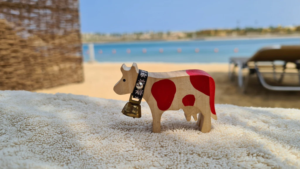 Figura de vaca de madera con manchas rojas y campana, colocada sobre una toalla clara, con playa de arena, agua azul y tumbonas al fondo.