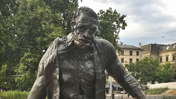 Escultura de bronce de una criatura humanoide con extremidades asimétricas, situada al aire libre sobre suelo rojo en un entorno urbano de Ginebra.