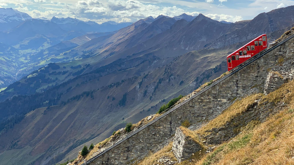 Vista de un funicular rojo del Niesen subiendo una pendiente empinada de montaña, con un valle y picos alpinos al fondo.