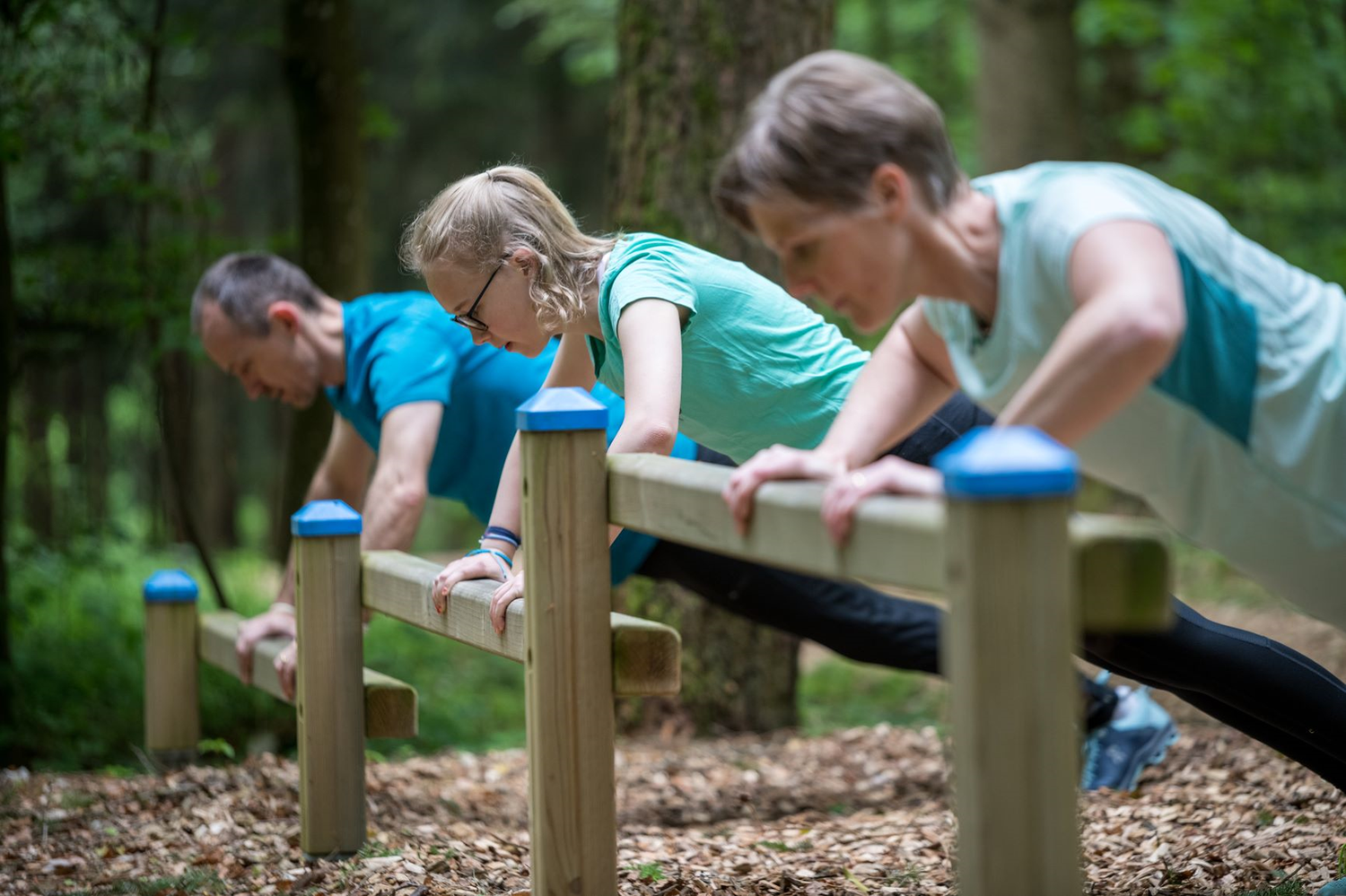 Une image montre une personne en tenue de sport portant une lourde bûche sur ses épaules. La personne se trouve à l'extérieur, dans une zone boisée, à côté d'un tas de bois.