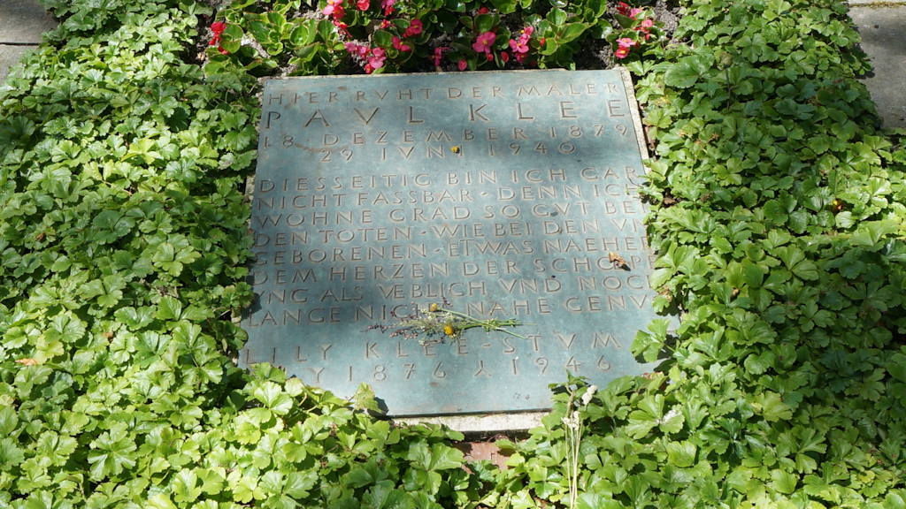 Tombe recouverte de verdure avec une plaque commémorative gravée et des fleurs colorées, située dans un cimetière arboré.