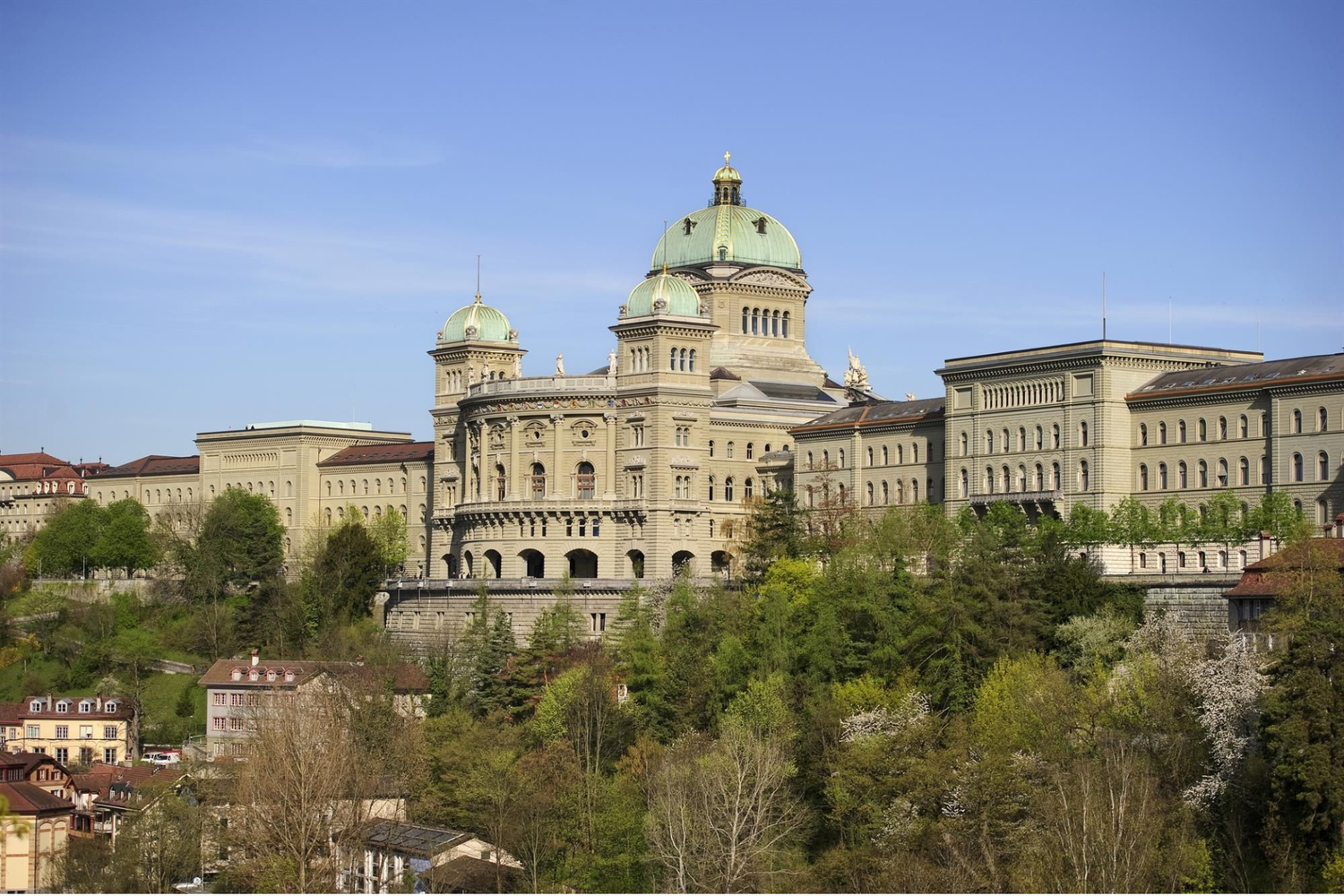 Vue extérieure du Palais fédéral suisse à Berne perché sur une terrasse rocheuse, montrant ses ailes symétriques et ses dômes verts au-dessus d’un talus boisé