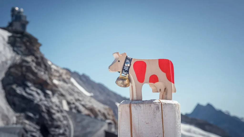 Figurine de vache en bois avec taches rouges et cloche, posée sur un poteau en bois, avec des montagnes enneigées et un ciel bleu en arrière-plan.