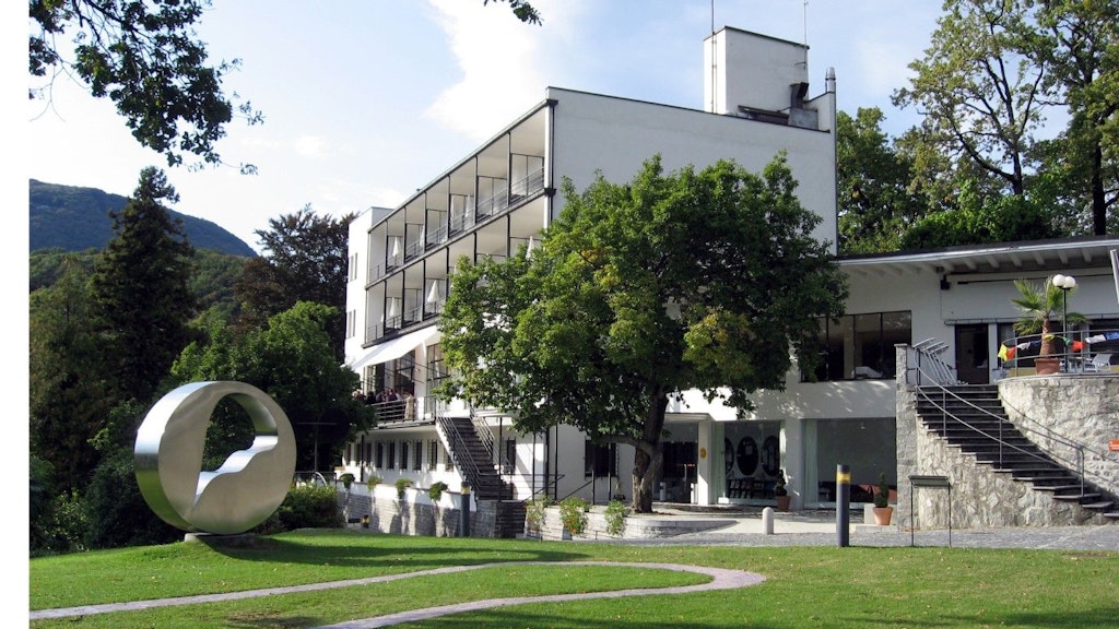 Bâtiment moderne de plusieurs étages avec balcons, entouré d’arbres, avec une sculpture circulaire en métal dans un jardin au premier plan.