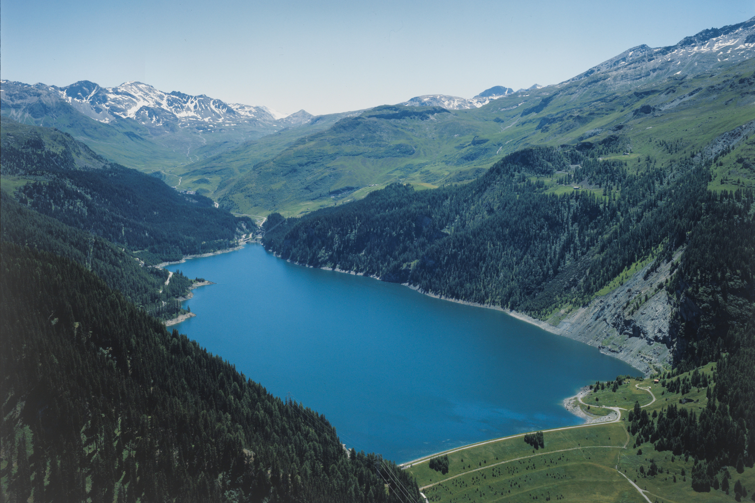 Vue aérienne du barrage de Marmorera, retenue d’eau turquoise entourée de forêts et de montagnes verdoyantes avec sommets enneigés au loin.