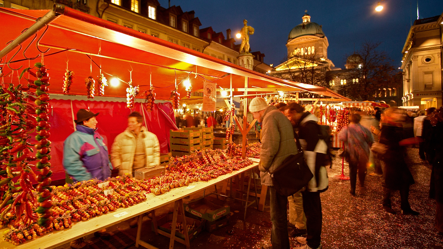 Marché nocturne animé avec étals illuminés vendant des guirlandes d'oignons, des visiteurs et bâtiments historiques visibles en arrière-plan.