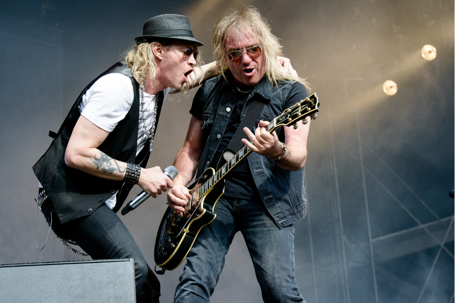 Deux musiciens sur scène, l’un chante dans un micro, l’autre joue de la guitare électrique, éclairés par des projecteurs dans une ambiance de concert.