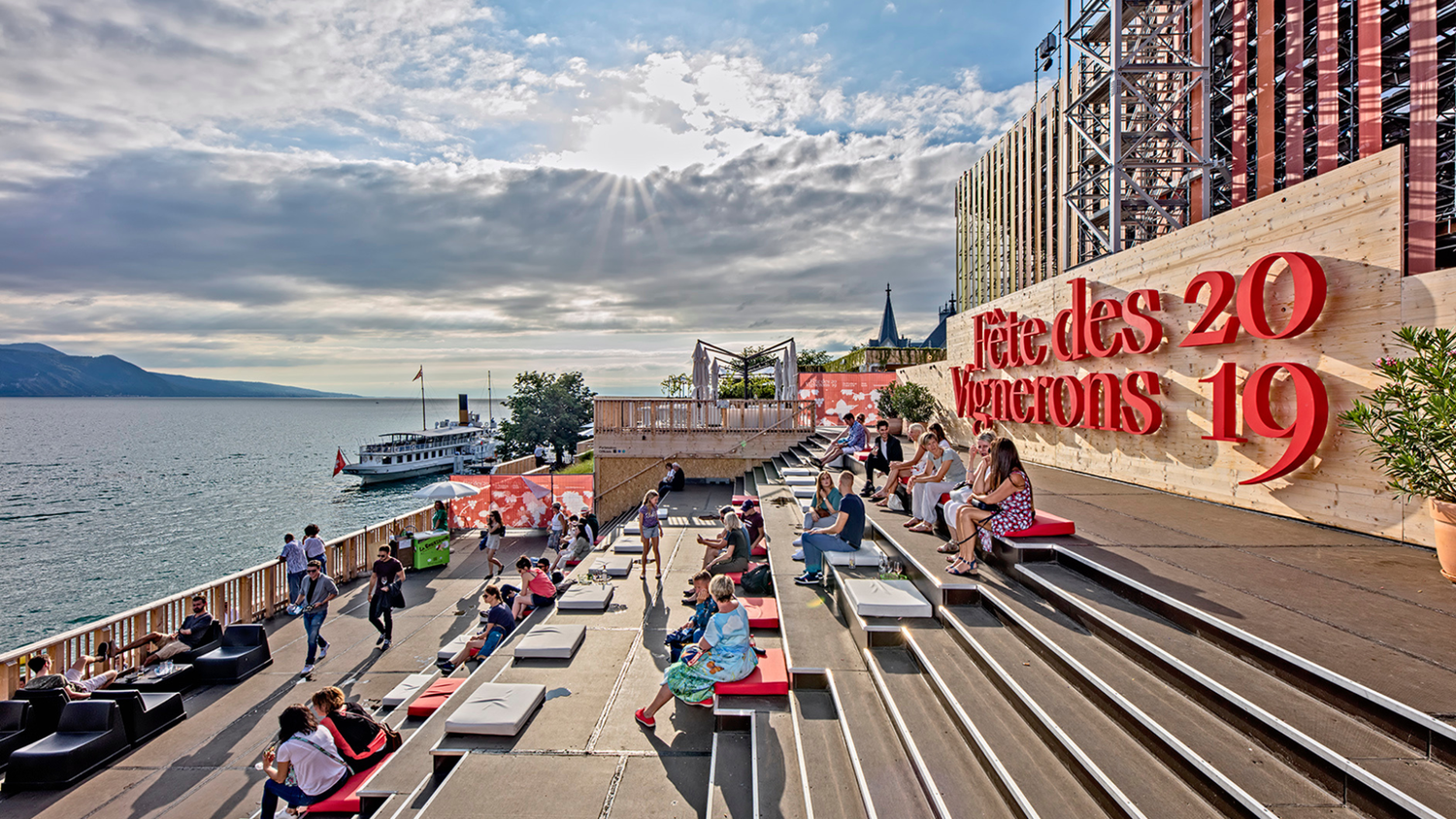Escaliers-terrasses de la Confrérie des Vignerons à la Fête des Vignerons 2019 à Vevey, surplombant le lac Léman, avec des visiteurs assis et un bateau en arrière-plan.