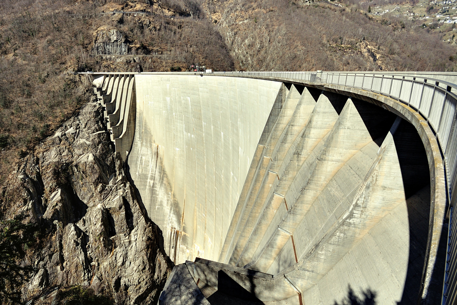 Vue du barrage de Contra, mur en béton incurvé inscrit entre des falaises rocheuses, passerelle avec rambarde en sommet.