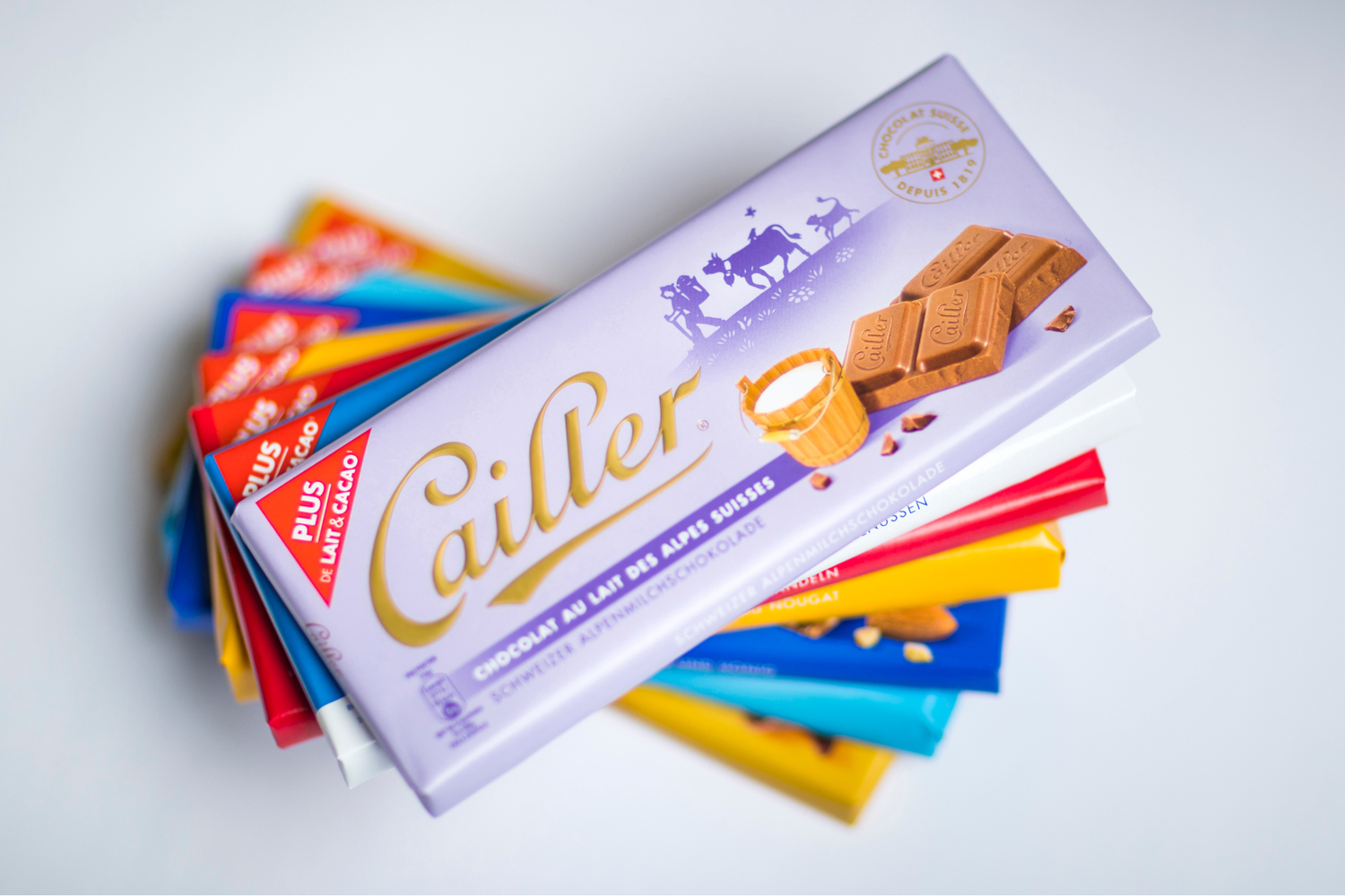 Paquet de chocolat Cailler empilé avec d’autres tablettes colorées, le dessus montre un emballage mauve illustrant des carrés de chocolat et un petit seau de lait.