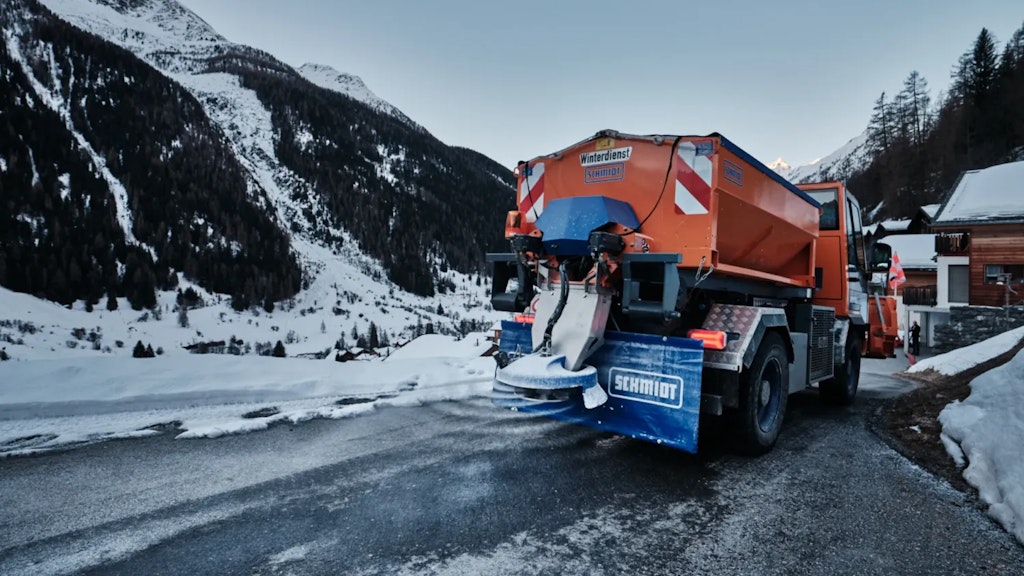 Camion de service hivernal orange équipé d’un épandeur circulant sur une route de montagne enneigée, avec villages et versants alpins en arrière-plan.