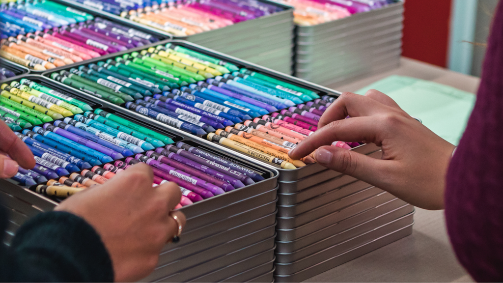 Mains choisissant des crayons pastel rangés par couleur dans des boîtes métalliques empilées. 