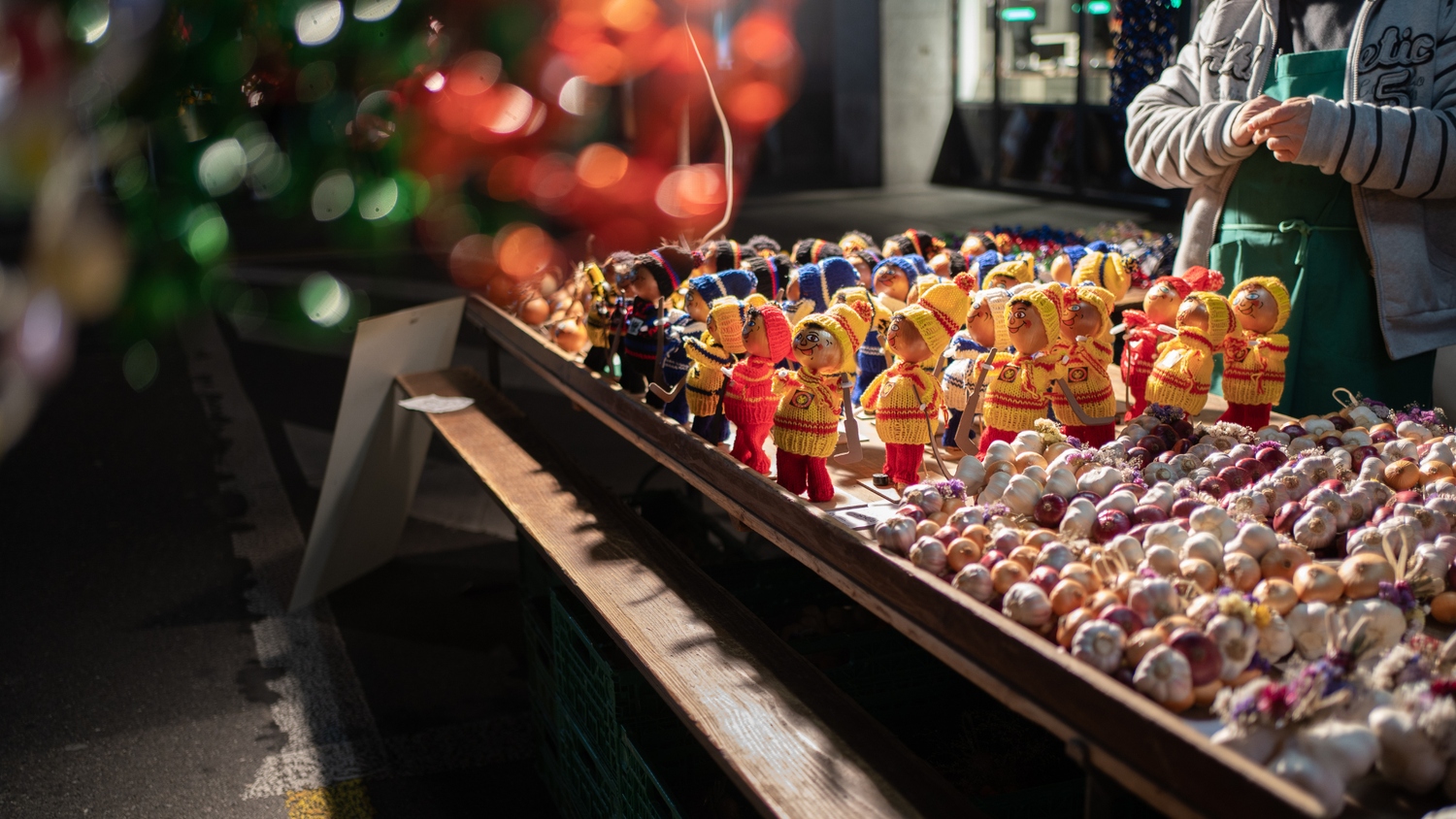 Stand de marché exposant des poupées colorées en tenue traditionnelle et des guirlandes d'oignons, avec un vendeur visible partiellement à droite.