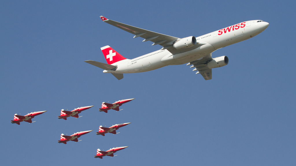 Avion de ligne Swiss en vol accompagné de six avions de chasse rouges et blancs sur fond de ciel bleu dégagé.