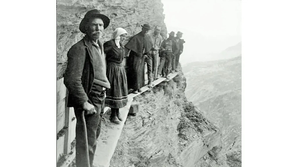 Photographie ancienne en noir et blanc montrant des hommes et une femme debout sur une étroite passerelle fixée à une paroi rocheuse en montagne.